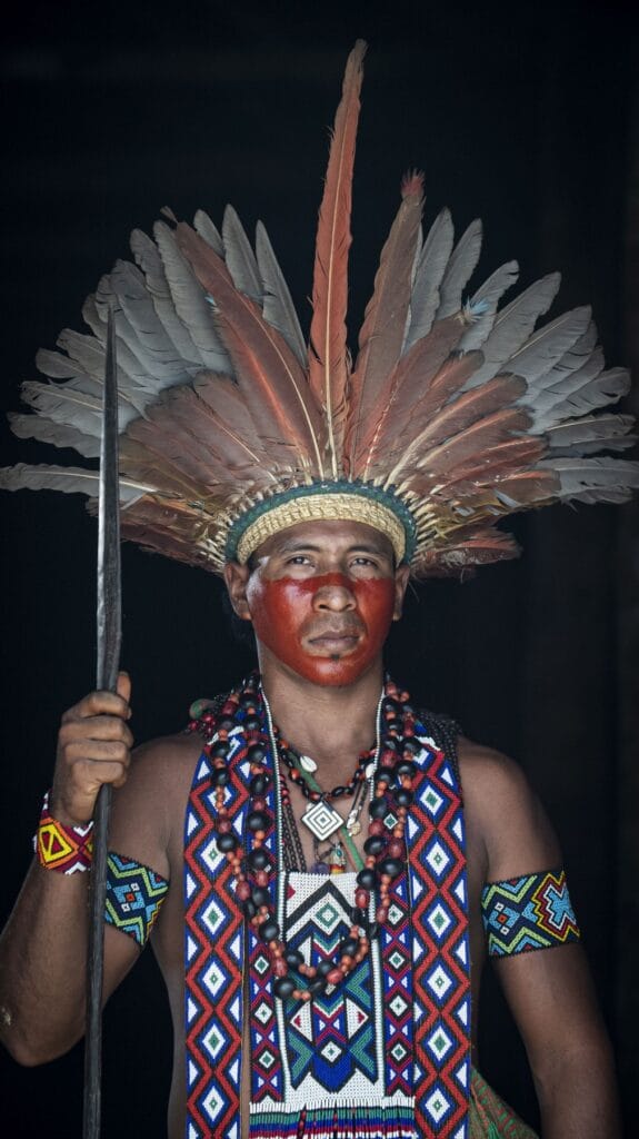 Hombre puyanawa con gran tocado de plumas rojizas, pintura roja en el rostro y chaleco de abalorios