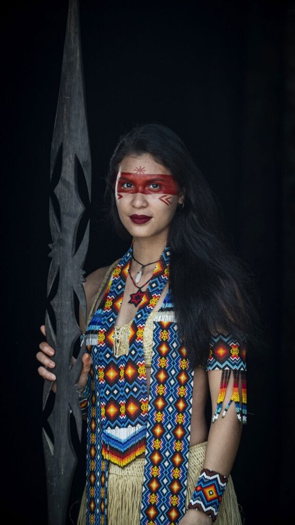 Mujer puyanawa con pintura facial roja, vestido de abalorios y lanza