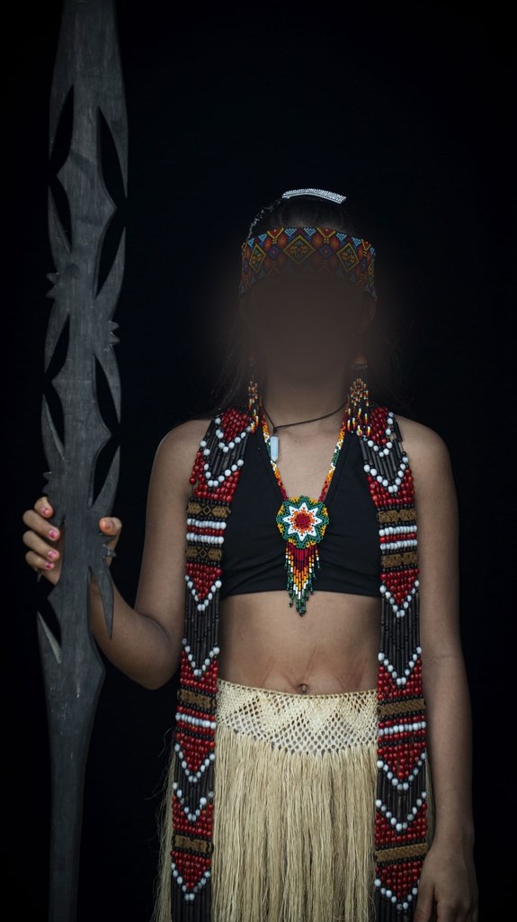 Chica puyanawa con cinta de abalorios en la frente, collar y falda de fibra, junto a una lanza