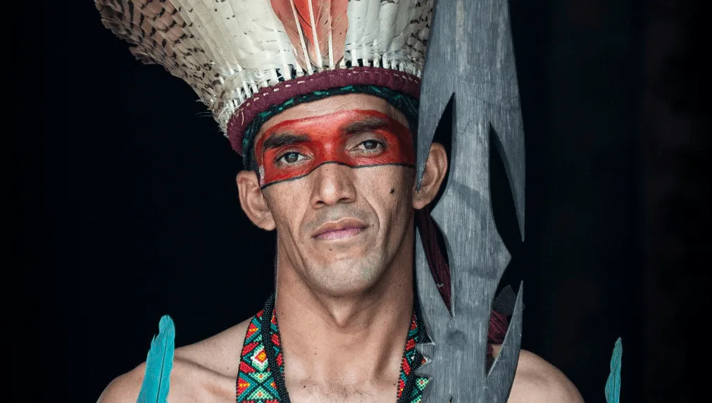 Retrato de un hombre puyanawa con tocado de plumas, pintura facial roja y una lanza