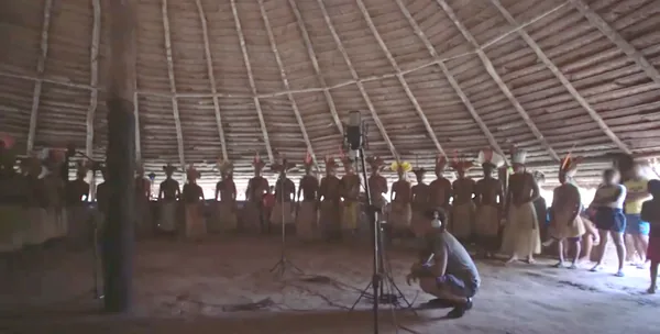 Cantores Puyanawa con tocados de plumas y faldas de paja, en fila dentro de la gran maloca, mientras una persona con auriculares, agachada junto al micrófono, controla la grabación de los cantos