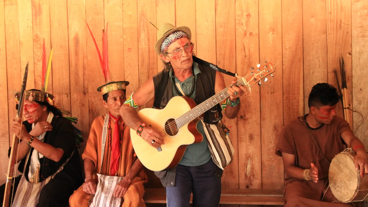 Txai Macedo tocando la guitarra sentado en un banco, junto a tres personas ashaninka con tocados y un tambor