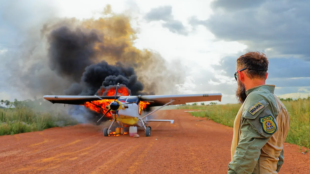 Una avioneta ardiendo en una pista de tierra en medio de la selva, con un agente ambiental de espaldas